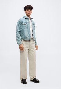 Νεαρός άνδρας που στέκεται φορώντας ανοιχτό μπλε τζιν μπουφάν, λευκό πουκάμισο, μπεζ παντελόνια και μαύρα παπούτσια, μπροστά σε απλό λευκό φόντο.