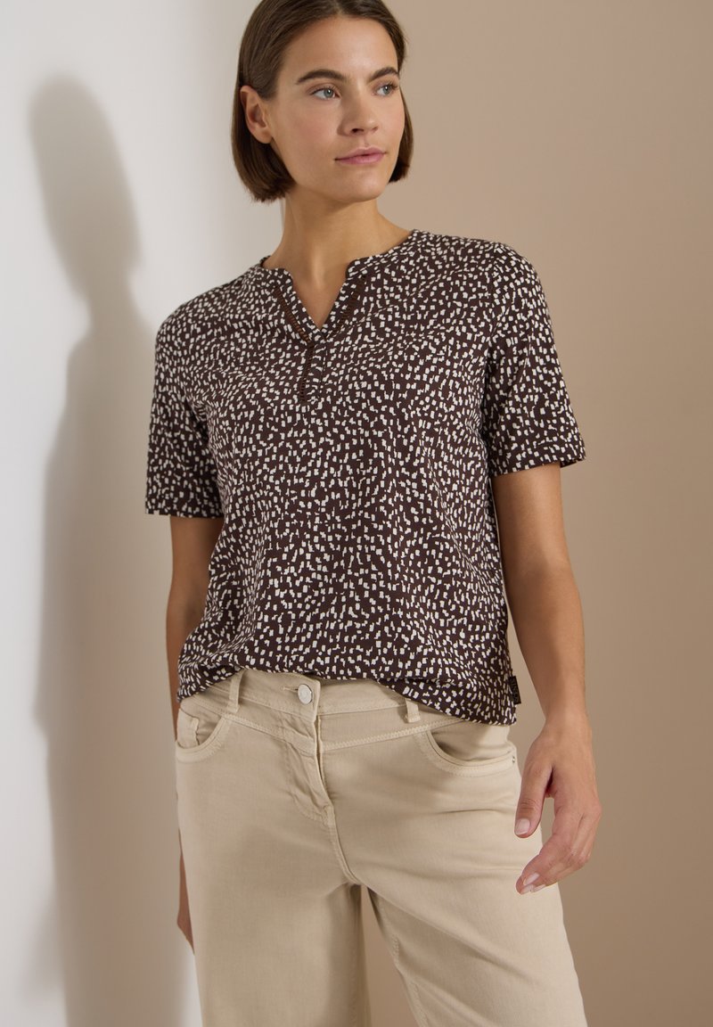 Femme portant un chemisier à manches courtes à motif noir et blanc et un pantalon beige, debout devant un mur bicolore beige et blanc.