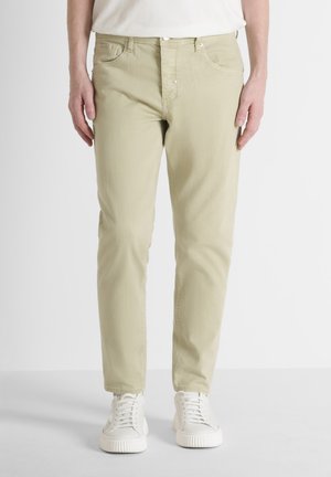 Man draagt beige slim-fit broek en witte sneakers, staat tegen een witte achtergrond met ontspannen handen langs zijn zij.