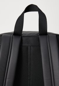 Black backpack top with a padded section, featuring mesh straps and a sturdy hanging loop. Textured surfaces with contrasting materials.