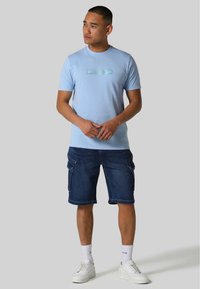 Lyseblå kortærmet t-shirt med et reflekterende logo, parret med knælange mørk denimshorts og hvide sneakers. Ensfarvet baggrund.