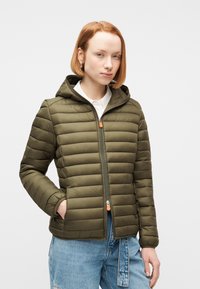 Γυναίκα με κοντά κόκκινα μαλλιά που φοράει ένα παλτό από πράσινο χακί καπιτονέ ύφασμα με κουκούλα, φερμουάρ κλειστό, και ανοιχτό μπλε τζιν, μπροστά σε απλό φόντο.