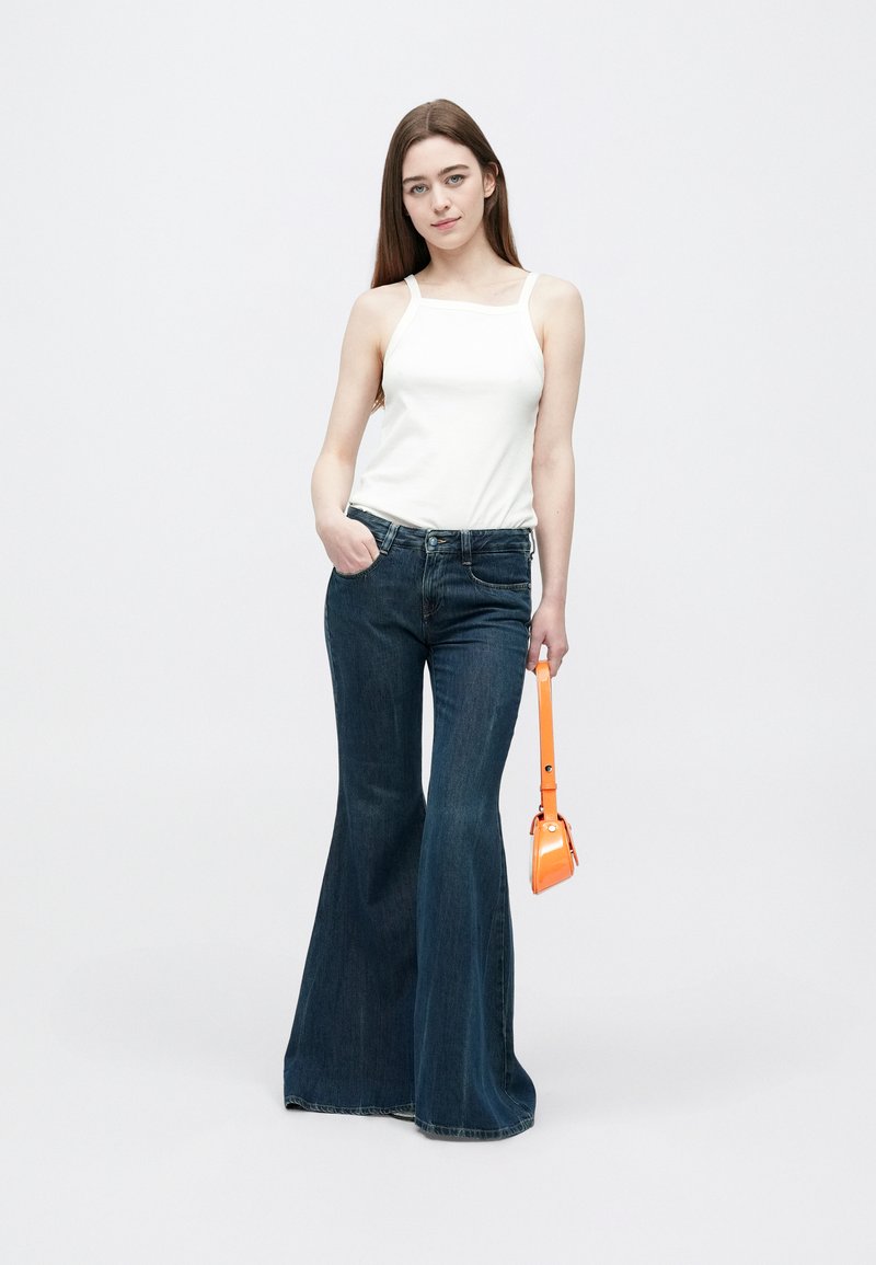 Young woman wearing white tank top and flared dark blue jeans holding a small orange handbag against a plain white background.