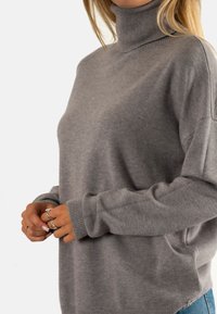 Suéter gris de cuello alto en material suave, con un corte holgado, cuello acanalado y aberturas laterales con un pequeño detalle de botón decorativo.