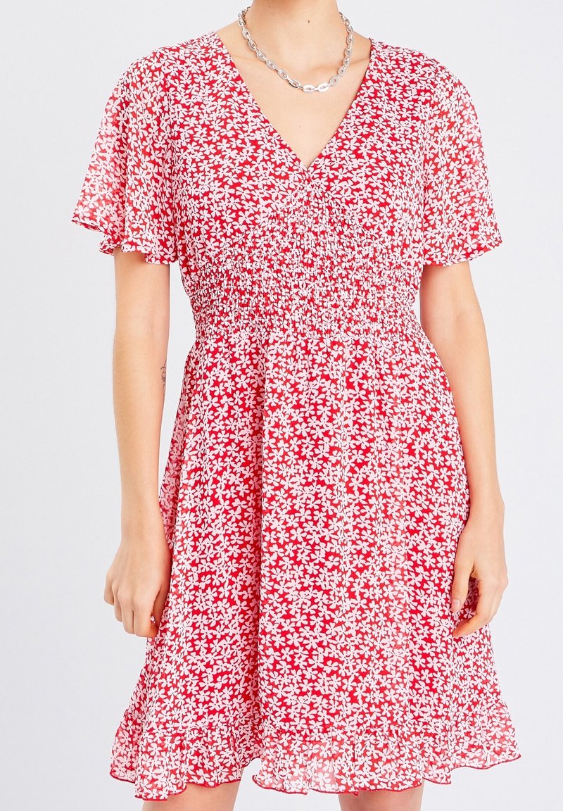 Robe florale rouge avec un décolleté en V, des manches courtes flottantes et une taille smockée. Tissu léger avec un ourlet évasé et un motif de fleurs blanches.