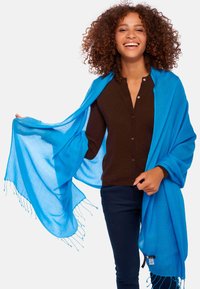 Écharpe légère bleu vif avec des bords frangés, drapée sur un haut marron foncé. Elle présente une texture douce et un design fluide.