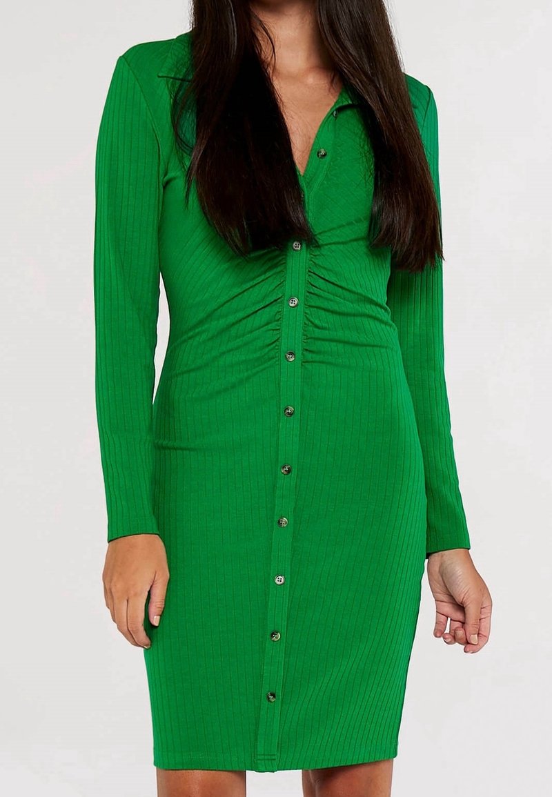 Femme portant une robe verte côtelée, ajustée, à manches longues, boutonnée, avec un léger froncé à la taille.