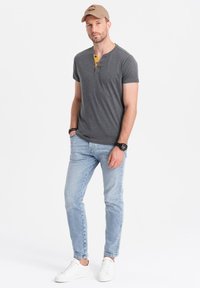 Camiseta de henley de manga corta gris con botones amarillos, combinada con jeans ajustados de color azul claro y zapatillas blancas. El modelo lleva una gorra beige.