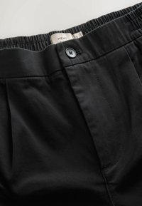 Fekete nadrág, amely textúrázott anyagból készült, rugalmas derékpánttal és gombos záródással, valamint oldalsó varrási részletekkel rendelkezik. Címke a derékpánton található.