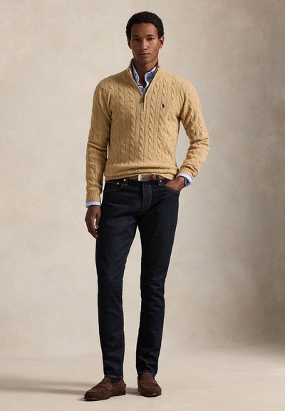 Ung mand står iført en beige kabelstriktrøje over en lyseblå skjorte, mørke jeans, brune sko og et ur med den ene hånd i lommen.