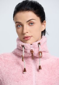 Chaqueta de forro polar de color rosa claro con cuello alto, presenta una cremallera frontal, cordones con detalles de madera y una superficie suave y texturizada.