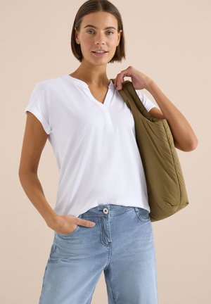 Femme aux cheveux courts bruns portant un haut blanc à manches courtes avec un col en V et un pantalon rayé bleu, portant un sac à bandoulière vert olive.