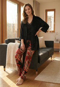 Vrouw in zwart topje en kamerjas met rode paisley broek staat op blote voeten, leunend op een grijze bank in een zonnige woonkamer met houten vloer.