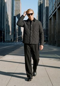 Homme blond portant des lunettes de soleil, une veste matelassée noire, un pantalon noir et des chaussures noires marche sur un trottoir en ville entre de grands immeubles.