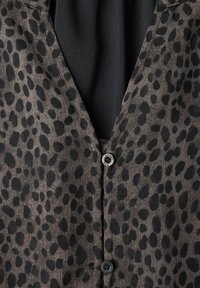 Chemise gris foncé à boutons avec des taches noires irrégulières et deux boutons visibles sur un fond en tissu noir.