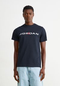 Melnas kokvilnas T-krekls ar "JORDAN" un sarkanu logo grafiku priekšā. Apaļš kakls, īsas piedurknes, apvienots ar gaiši ziliem džinsu šortiem.