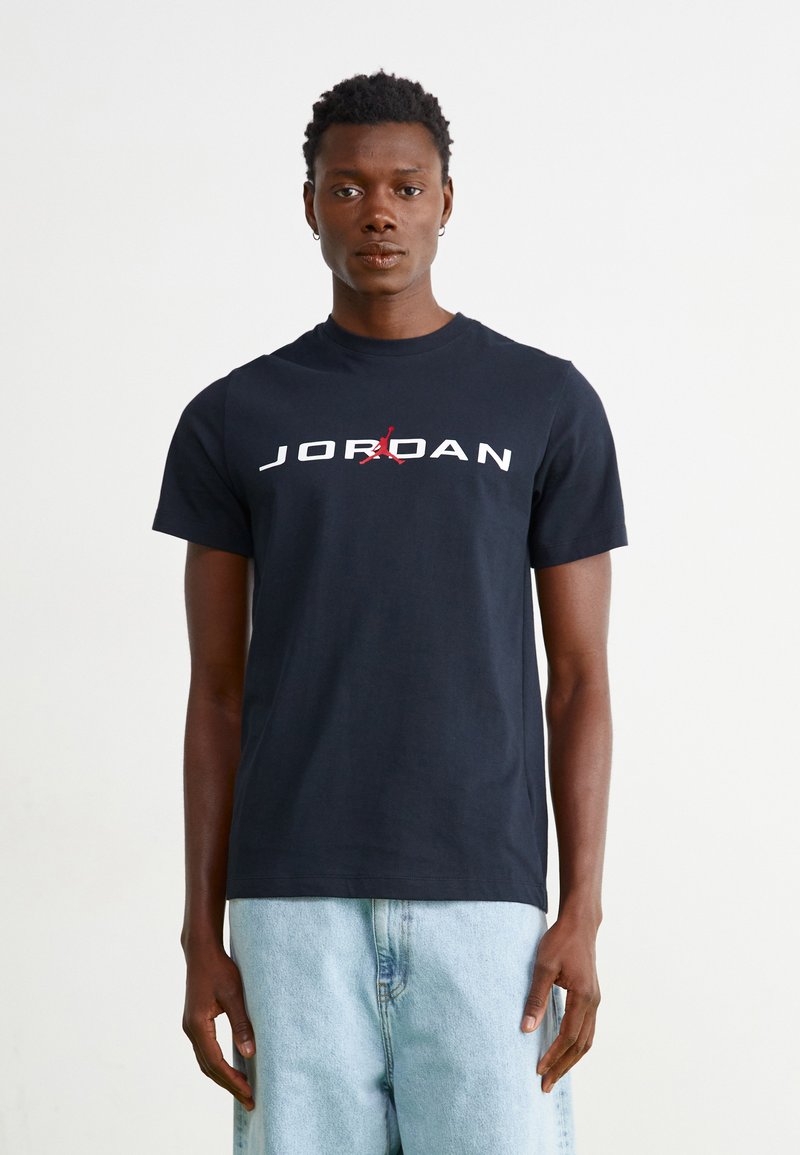 Melnas kokvilnas T-krekls ar "JORDAN" un sarkanu logo grafiku priekšā. Apaļš kakls, īsas piedurknes, apvienots ar gaiši ziliem džinsu šortiem.