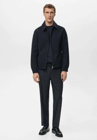 Granatowa kurtka na zamek wykonana z teksturowanej tkaniny, z charakterystycznym ostro zakończonym kołnierzem. Nosi się ją na ciemnym swetrze, w zestawieniu z dopasowanymi ciemnymi spodniami i czarnymi butami.