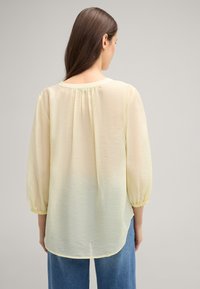 Blusa leggera gialla, trasparente, con maniche a sbuffo, scollatura rotonda e dettaglio arricciato sul retro. Tessuto liscio, scende sotto i fianchi.