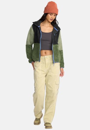 Veste en polaire verte et noire avec des fermetures éclair bleues, associée à un pantalon cargo crème et un bonnet en tricot couleur rouille.