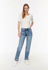 Biała koszulka z krótkim rękawem z logo, zestawiona z jasnoniebieskimi spodniami typu straight-leg. Model nosi białe sneakersy, stoi na jednolitym tle.