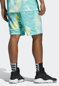 Pantalones cortos de malla de color turquesa claro con un patrón de tie-dye en amarillo y blanco, que cuentan con franjas laterales y un ajuste relajado, combinados con zapatillas deportivas negras.