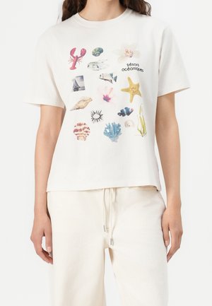 Vrouw draagt een roomwit T-shirt met kleurrijke prints in oceaanstijl, waaronder een kreeft, zeester, schelp, vis, koraal en de tekst "trésors océaniques."