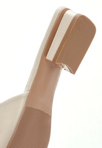 Beige hoge hak schoen met een glad leren bovenwerk, vierkante hak en een textuur rubberen zool. Bevat een contrasterende witte accent nabij de hak.