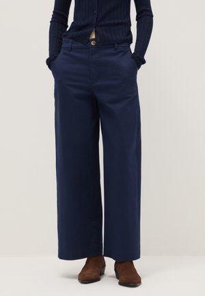 Persona que lleva pantalones anchos azul marino, cárdigan negro acanalado y botines marrones, de pie con las manos en los bolsillos sobre un fondo liso.