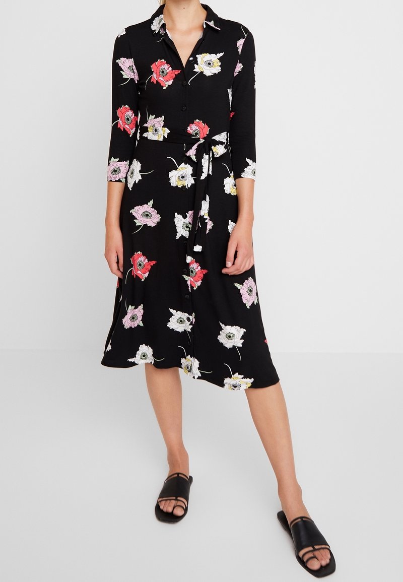 Svart klänning med blommönster i rosa, rött och vitt. Långa ärmar, krage, knappfront och detalj med midjebälte. Svarta sandaler.
