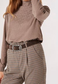 Gestrickter brauner Pullover mit strukturiertem Muster am Ausschnitt, kombiniert mit hoch taillierten Hosen im Hahnentrittmuster und einem braunen Gürtel mit verziertem silbernem Schnallenverschluss.