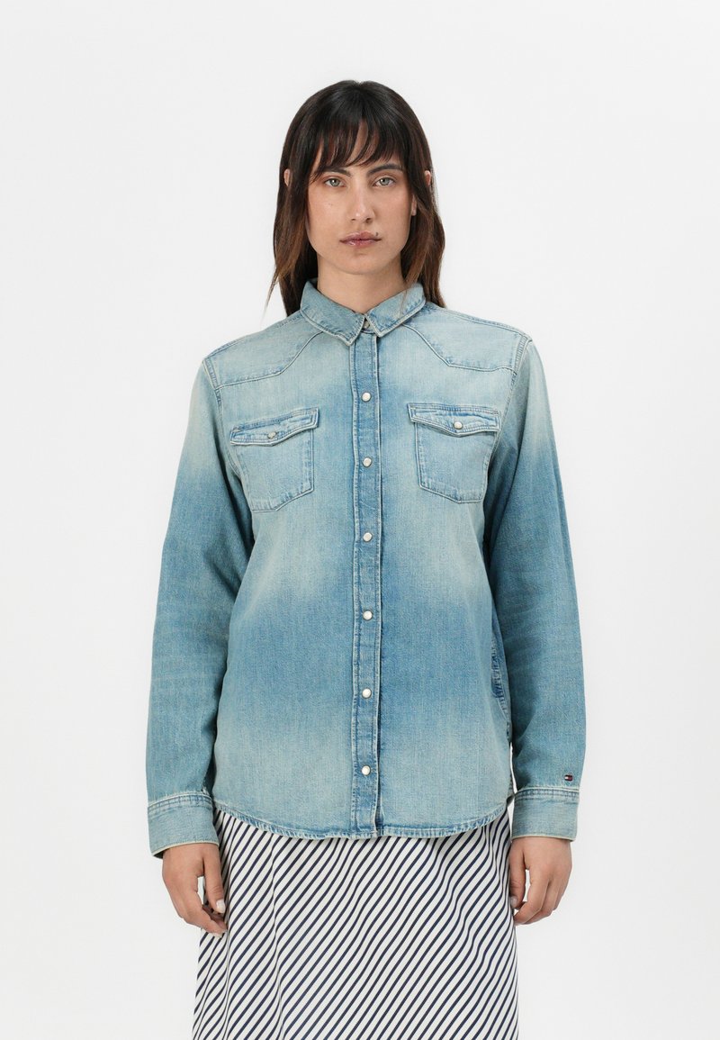 Camisa de denim azul clara com colarinho, dois bolsos frontais e fechos de botão. Apresenta uma textura desbotada e detalhes cosidos.