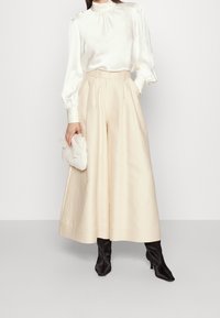 Donna che indossa una blusa di seta crema con maniche a sbuffo, pantaloni larghi di colore crema, stivaletti neri a punta, mentre tiene una piccola pochette crema.