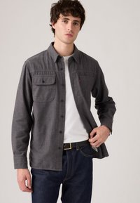 Grijze overhemd met knopen, twee borstzakken, lange mouwen en een relaxte pasvorm, gecombineerd met blauwe jeans en een zwarte riem.