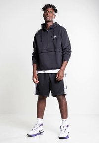 Hoodie preta com fecho de correr e cordões, combinada com calções pretos com detalhes laterais. Usado com sapatilhas desportivas brancas e roxas.