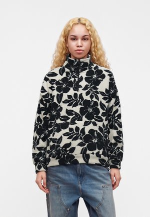 Ung kvinde med krøllet blondt hår iført en sort-hvid blomsterfleece-pullover og blå jeans, stående mod en ensfarvet baggrund.