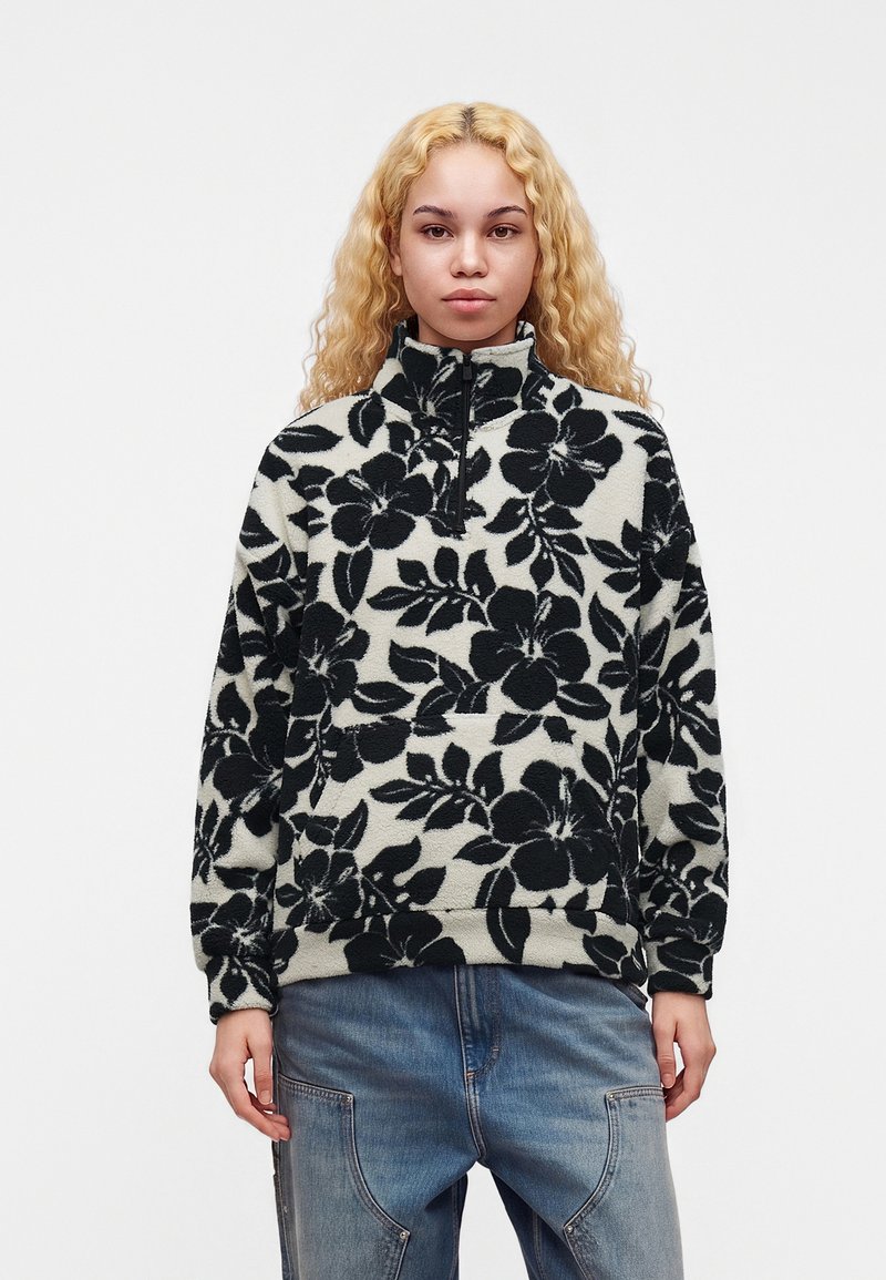 Jeune femme aux cheveux blonds bouclés portant un pull polaire à fleurs noir et blanc et un jean bleu, se tenant devant un fond uni.