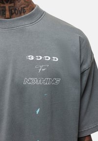 Szara, bawełniana, oversize t-shirt z napisem "GOOD For NOTHING" w różnych czcionkach, z jasnoniebieskimi plamami farby i okrągłym dekoltem.
