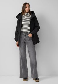 Zwarte waterdichte jas met een capuchon, gedragen over een gestreepte coltrui. Grijze wijde jeans met zichtbare stiksels, gecombineerd met zwarte laarzen.