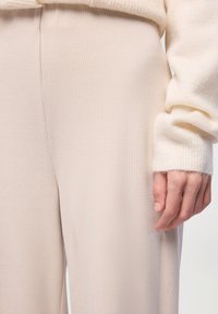 Zbliżenie na prawą dłoń osoby obok beżowych spodni o teksturze oraz miękkiej, kremowej bluzki z długim rękawem.