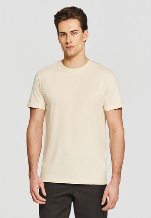 Młody mężczyzna z krótkimi brązowymi włosami, ubrany w jednolity beżowy t-shirt z krótkim rękawem i czarne spodnie na jasnoszarym tle.