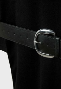 Čierny kožený opasok s lesklou striebornou oválnou sponou zapnutý okolo čierneho semišového materiálu.