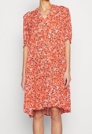 Frau trägt ein locker sitzendes orangenes Kleid mit kleinem weißem und schwarzem Blumenmuster und ellbogenlangen Ärmeln.