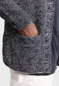 Cardigan texturé noir et blanc avec un motif tricoté, présentant une poche avant et des poignets côtelés. Le tissu semble doux et chaud.