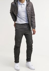 Vadderad grå jacka med huva, kombinerad med en ljusgrå pikétröja och mörkgrå byxor. Vita sneakers fulländar outfiten.