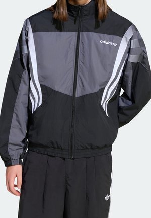 Personne portant une veste de survêtement Adidas noire et grise avec des bandes blanches et un pantalon de survêtement Adidas noir avec logo sur la jambe gauche.