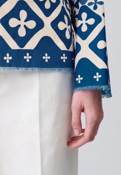 Top bluetto con motivo geometrico in cotone e maniche sfrangiate, abbinato a una gonna in denim bianco texturizzato. Mano appoggiata sulla gonna.