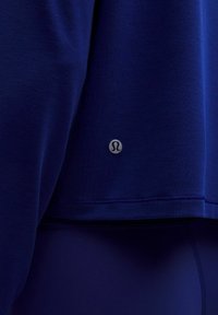 Tricou sportiv de lungime medie, de culoare albastră închisă, cu un tiv rotunjit și o etichetă cu logo argintie pe partea inferioară. Textură de material moale și neted.