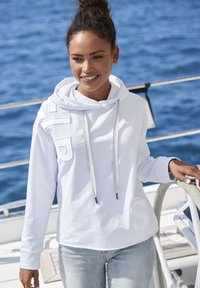 Weißer Kapuzenpullover mit grauen Kordeln, auf der Vorderseite mit strukturierten Buchstaben. Getragen mit hellblauen Jeans vor einem blauen Hintergrund.