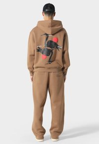 Brun hættetrøje og sweatpants sæt. Hættetrøjen har en sort stork grafik og røde detaljer på ryggen. Blødt, tekstureret stof, afslappet pasform.